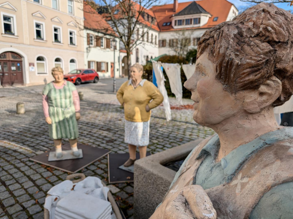 Drei bemalte Figuren auf einem gepflasterten Platz in einer Altstadt.